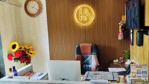A reception desk with a laptop, papers, and a vase of sunflowers. A wooden panel with a neon sign and a clock are visible in the background. - DhamStay Hotel Aradhya Near Beach Puri  (Puri)