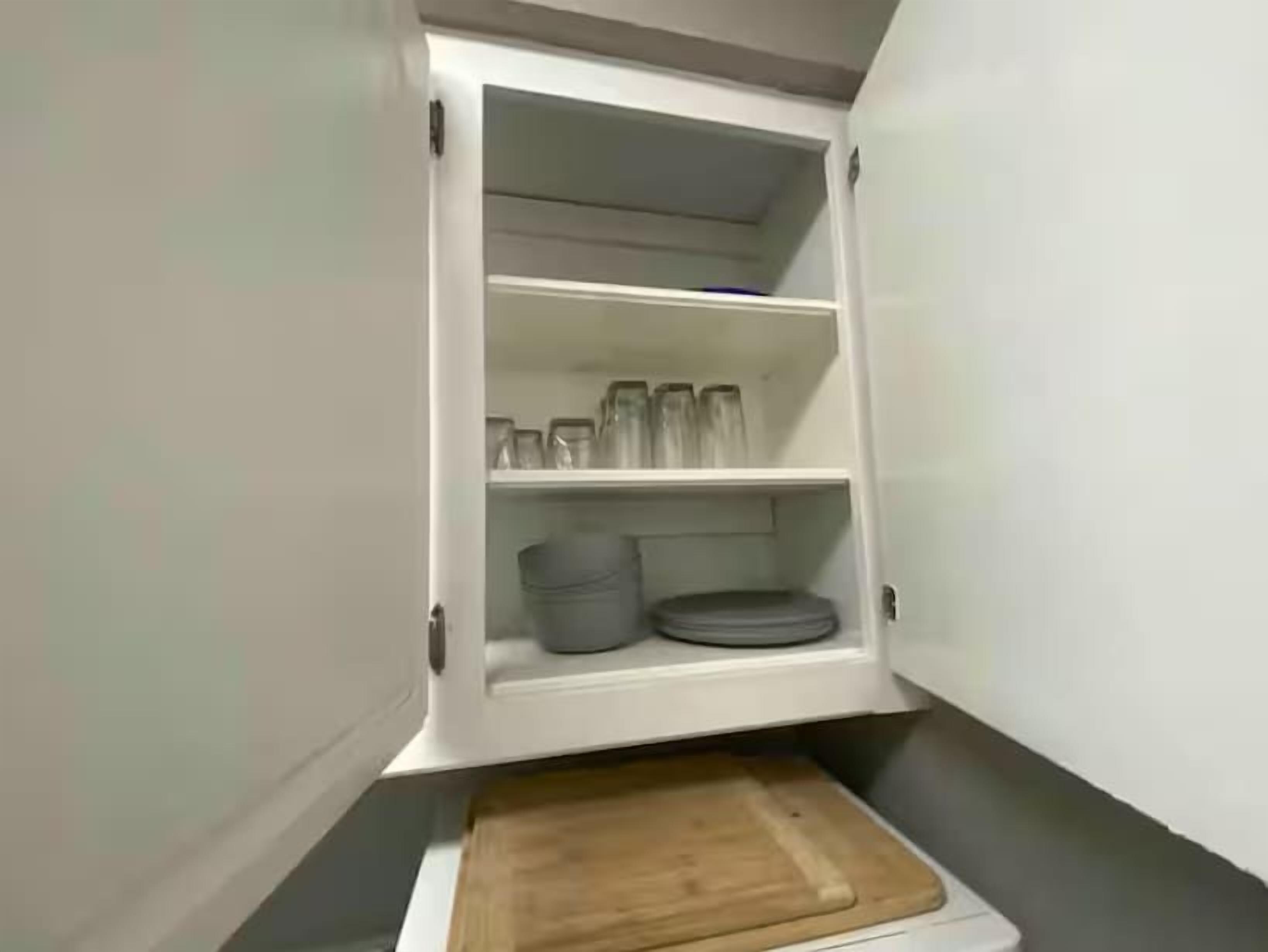 A kitchen with a wooden cutting board on the counter and a white cabinet with shelves containing glassware and plates.