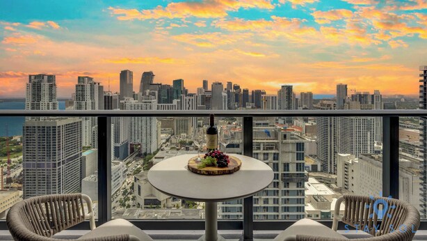 A balcony with a round table, two wicker chairs, a bottle of wine, and a fruit platter. - Downtown 2BR Oasis: Pool, Gym, Balcony Views (Miami)