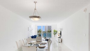 A modern living room with a glass dining table, white sofas, and a large window offering a view of a pool and buildings. - 2BR 2BTH - Apartment  - Free Parking (Sunny Isles Beach)