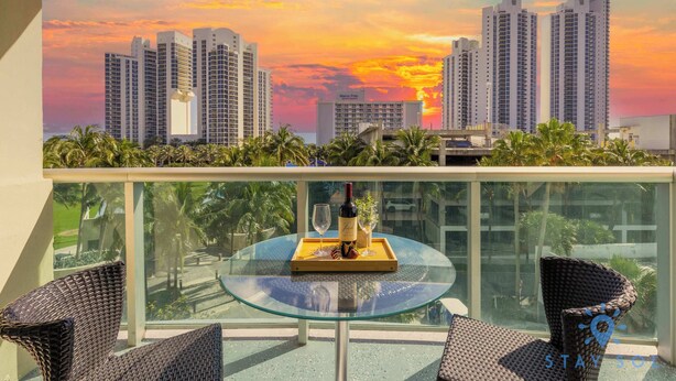A balcony with a glass table, two wicker chairs, a bottle of wine, and a tray with a glass. - 2BR 2BTH - Apartment  - Free Parking (Sunny Isles Beach)