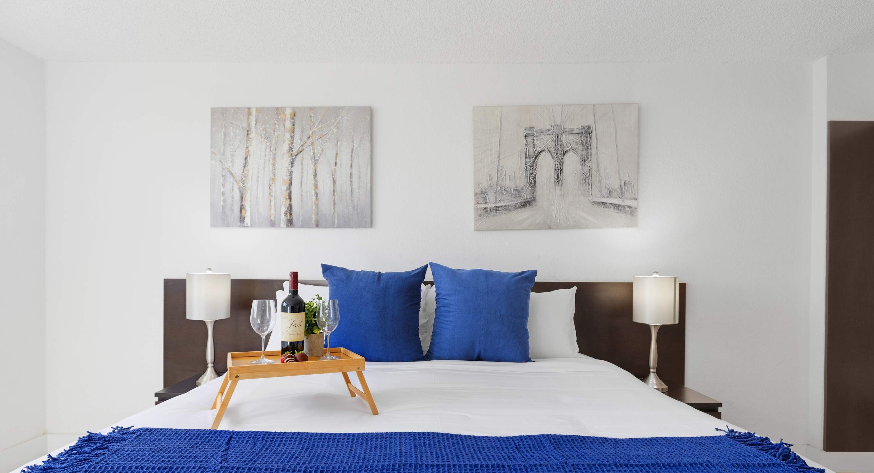 A hotel room with a bed, two blue pillows, a wooden bedside table with a tray holding a bottle and glasses, and two wall-mounted artworks depicting a forest and a bridge.