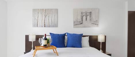 A hotel room with a bed, two blue pillows, a wooden bedside table with a tray holding a bottle and glasses, and two wall-mounted artworks depicting a forest and a bridge.
