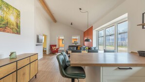 Une cuisine spacieuse avec un îlot central en bois, un coin repas avec une table et des chaises, et de grandes fenêtres offrant une vue sur l’extérieur.