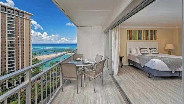 A balcony with a table and chairs, a bed with a view of the ocean, and a building in the background. - @ Marbella Lane - Ocean Marina View | Ilikai Hotel (Honolulu)
