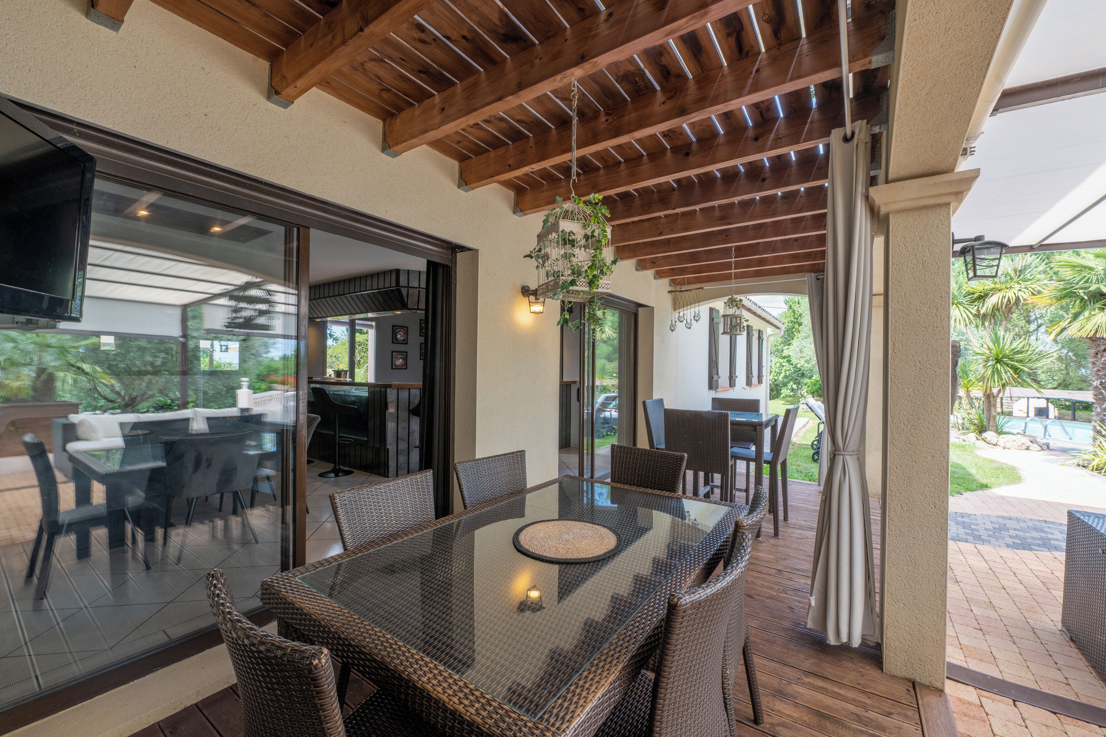 A covered patio with a glass table and wicker chairs, overlooking a backyard with a barbecue area and outdoor seating.