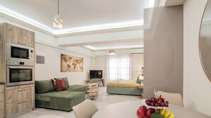 A modern living room with a green sofa, a dining table with fruit, and a kitchen area with built-in appliances.