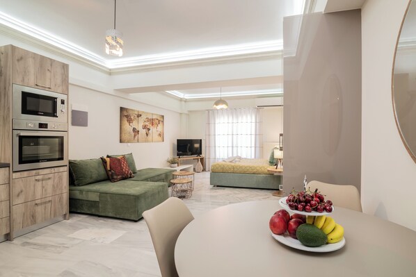 A modern living room with a green sofa, a dining table with fruit, and a kitchen area with built-in appliances. - ExAnimo Luxury Apt 2 (Zakinthos)