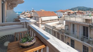 A balcony with a wooden table and a wicker basket of plants, overlooking a coastal town with terracotta-roofed buildings and a clear blue sky.