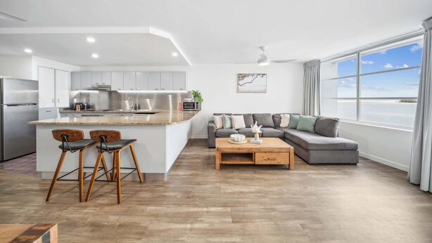 Una cocina moderna con una isla central, un área de comedor con sofá y mesa de centro, y un ventanal con vistas.