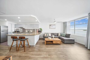 A modern kitchen with a central island, a dining area with a sofa and coffee table, and a large window with a view. - Pumicestone Passage Waterfront Escape (Caloundra Region)