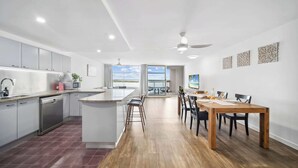 Una cocina moderna con una isla central, un área de comedor con mesa y sillas de madera, y vistas a través de la puerta de cristal.
