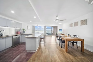A modern kitchen with a central island, a dining area with a wooden table and chairs, and a view through the glass door.