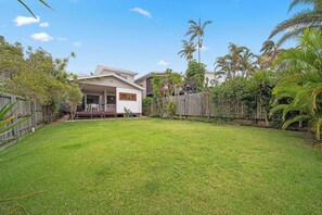 Un patio trasero residencial con una casa, cerco de madera y vegetación tropical.