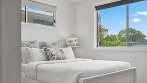 Una cama blanca con almohadas a rayas, una lámpara de noche y una ventana con vistas a árboles.