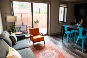 A living room with a sofa, a coffee table, and a TV. There is a sliding glass door leading to a patio with outdoor furniture.