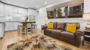 A modern kitchen with white cabinets, a dining area with a brown leather couch, and a large window with a view. - Capri 001 & 002- TWO Condos Downtown Traverse City (Traverse City)