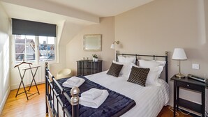 A neatly made bed with pillows and a blanket, a bedside table with a lamp, a small dresser, and a window with a view of buildings.