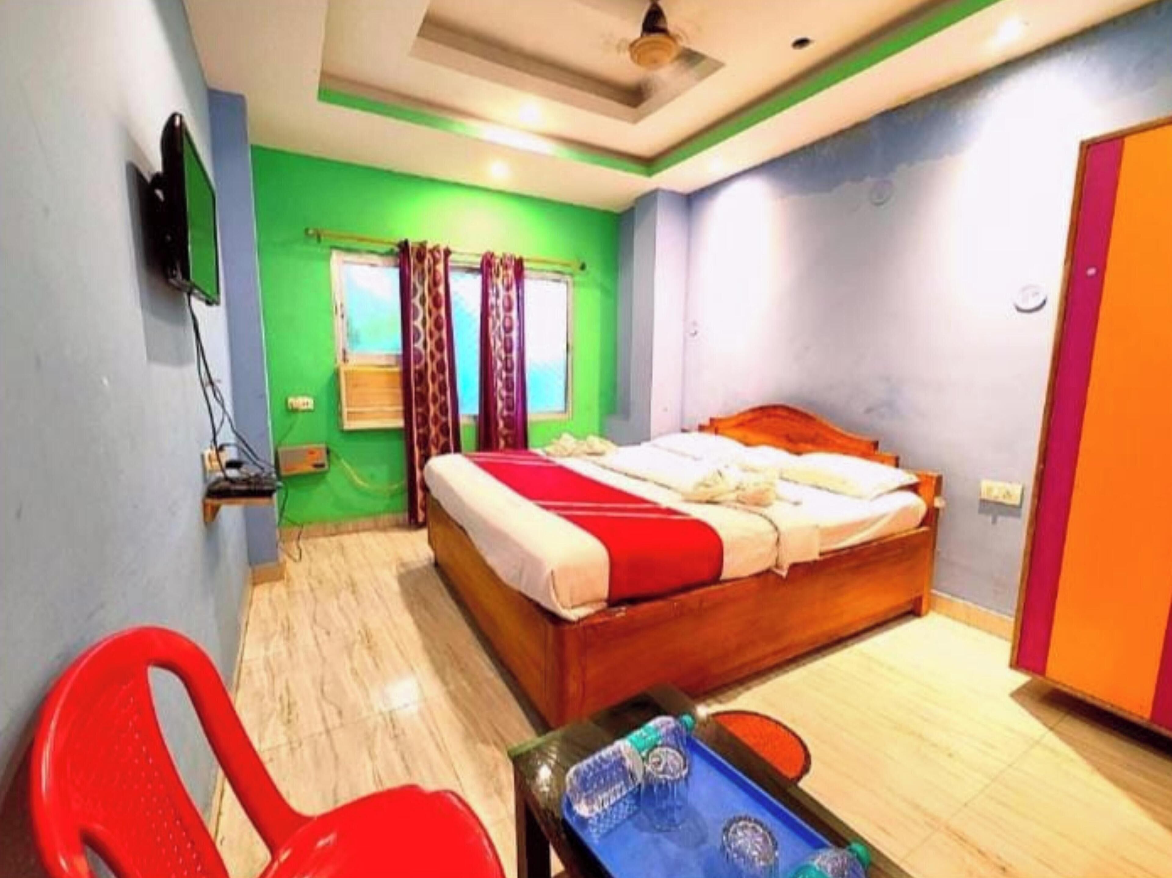 A room with a bed, a TV, a window with curtains, a chair, and a table with bottles.