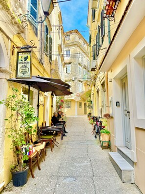 A narrow street with buildings on both sides, outdoor seating at a café, and a bicycle parked on the right. - Victoria's Old Town Gem - Cozy, Renovated Apartment in Corfu (Kerkira)