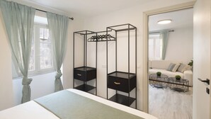Un dormitorio con cama, dos mesitas de noche negras con cajones, una ventana con cortinas ligeras y vistas a una sala de estar con sofá y dos mesas redondas.