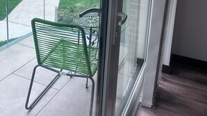 A balcony with a glass door, a green metal chair, and a paved pathway leading to a building with a brick facade.