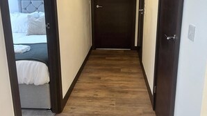 A narrow hallway with wooden flooring, a bed with white bedding, and a closed door at the end.