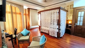 Quarto de hotel com piso de madeira, cama de dossel, cadeiras de vime e uma televisão.