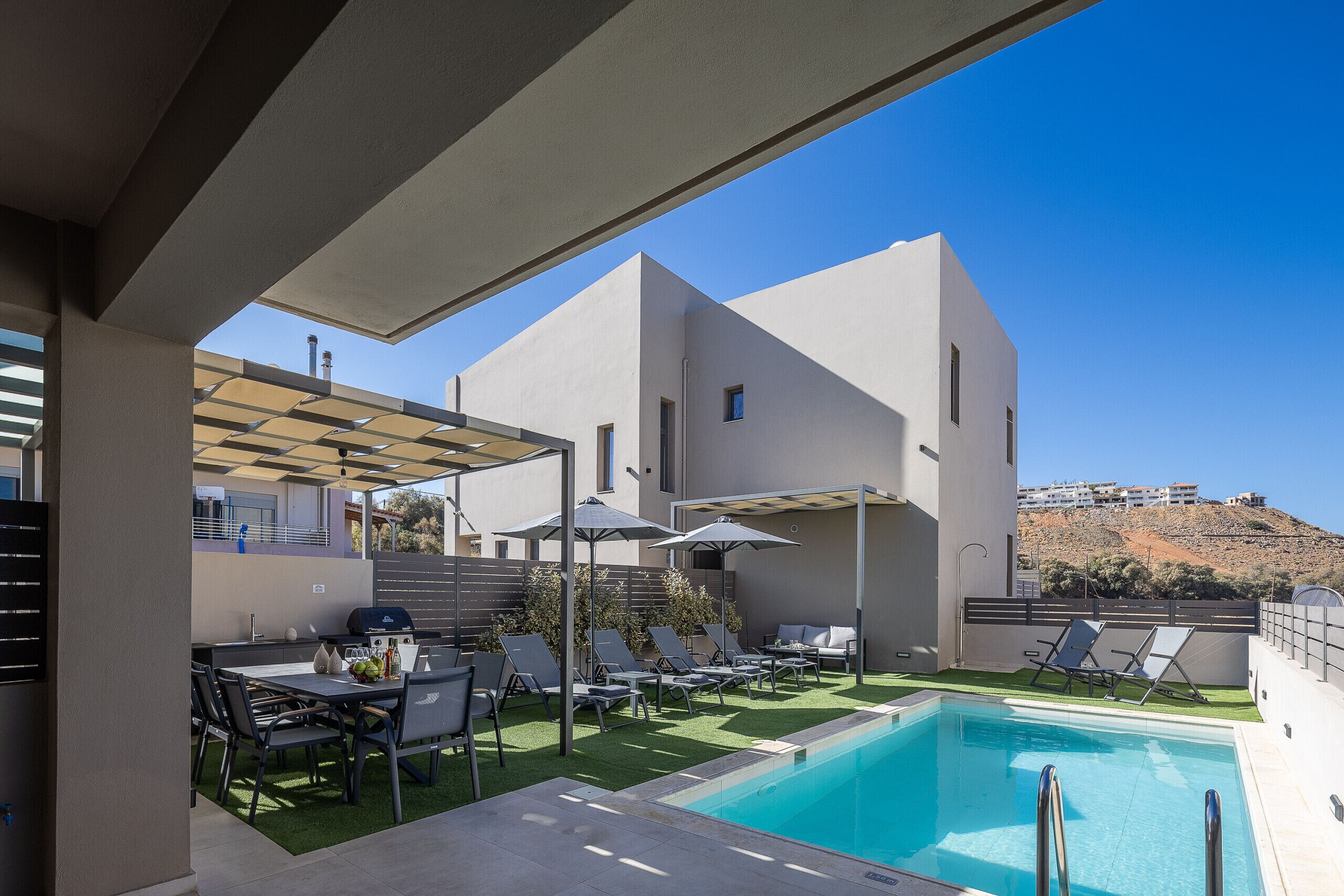 A modern outdoor pool area with a clear blue sky, a pool, lounge chairs, and a barbecue setup.