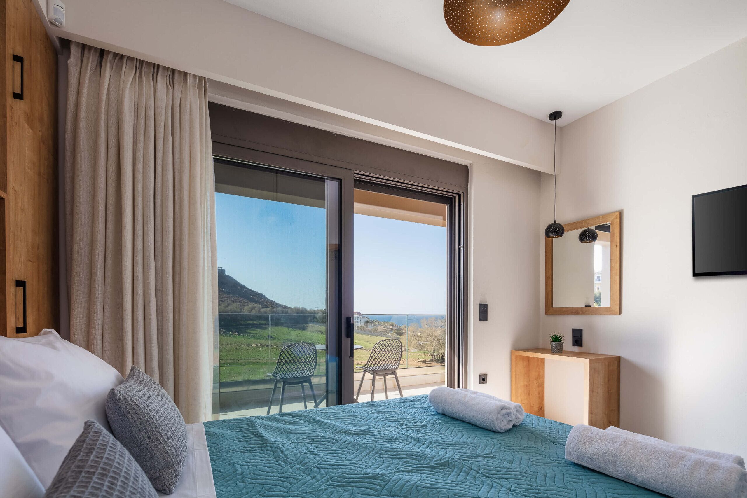 A bedroom with a large bed, a view of the outdoors, a wooden bedside table, and a mirror on the wall.