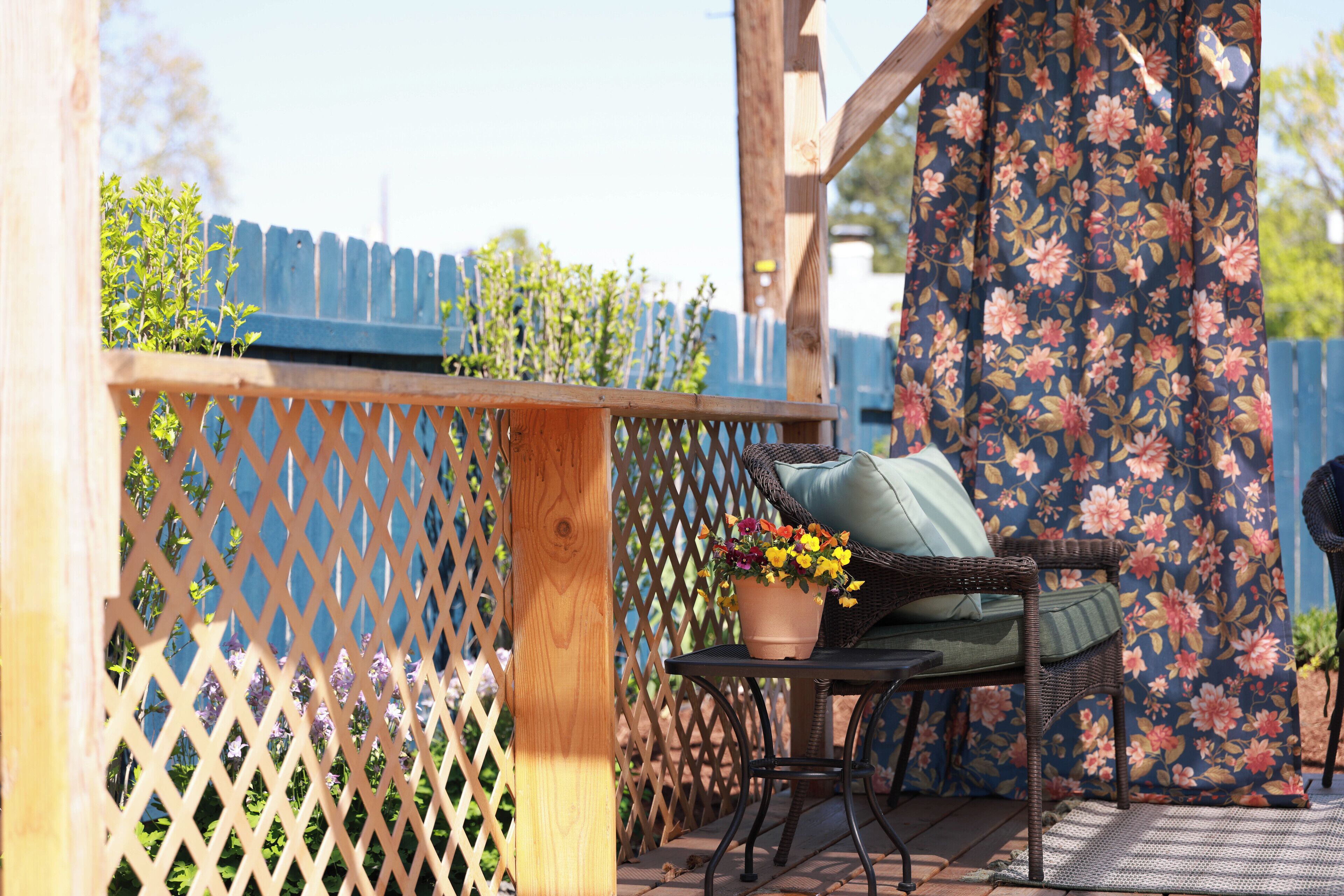 Eine Holzterrasse mit Gitterzaun, ein Korbstuhl mit Kissen und ein Tisch mit einem Topfpflanzen.