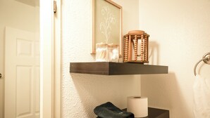 Ein Badezimmer mit einem hölzernen Ablagebrett, auf dem eine Kerze, ein Toilettenpapierrollen und ein gefaltetes Handtuch liegen.