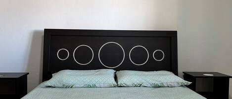 Una cama bien hecha con una colcha estampada y almohadas, un cuadro colgado en la pared que muestra un muelle, y una mesita de noche sencilla.