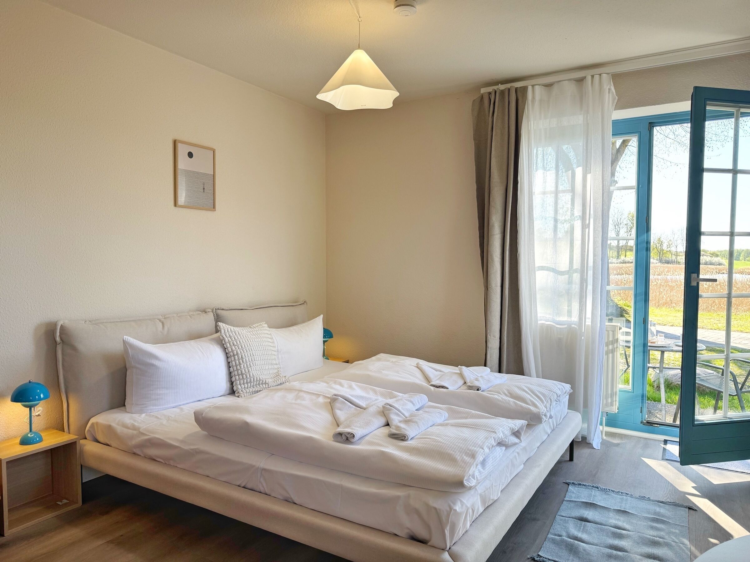 A neatly made bed with white linens and towels, a bedside lamp, and a view of the outdoors through a large window with curtains.