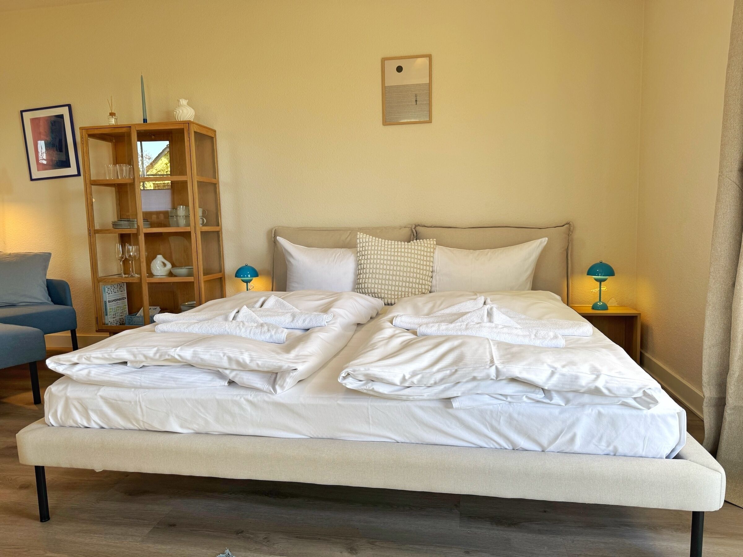 Ein ordentlich bezogenes Bett mit weißen Leinen, ein Nachttisch aus Holz mit einer Lampe, ein blauer Sessel und ein Bücherregal an der Wand.