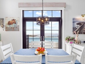 A dining area with a table set for two, a view of the sea through large windows, and a hanging light fixture. - Ocean Mist Cottage (Carna)