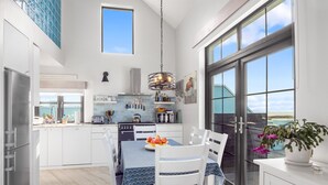 A modern kitchen with white cabinets, a dining table with a blue tablecloth, and a large window overlooking a scenic view. - Ocean Mist Luxurious Seaside Haven (Carna)