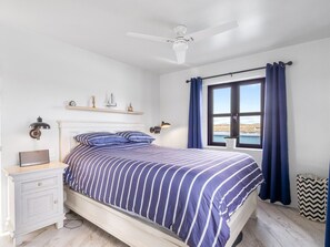 A bedroom with a bed, bedside table, lamp, and a window with curtains. - Ocean Mist Cottage (Carna)