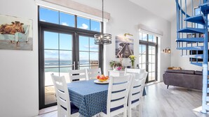 A dining area with a table set for two, a blue patterned tablecloth, white chairs, and a view of the ocean through large windows. - Ocean Mist Luxurious Seaside Haven (Carna)