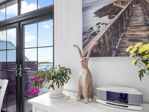 A room with a large window, a white desk, a decorative rabbit figurine, a potted plant with pink flowers, and a speaker. - Ocean Mist Cottage (Carna)