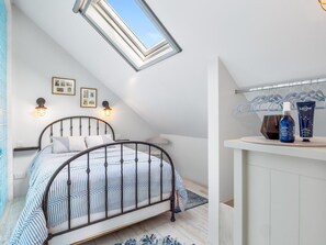 A bedroom with a sloped ceiling, a bed with a metal frame, a skylight, wall-mounted lights, a bedside table, and a clothing rack with items. - Ocean Mist Cottage (Carna)