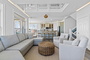 Een moderne woonkamer met een hoekbank, een rond rieten pouf en een eethoek, met een keuken op de achtergrond.