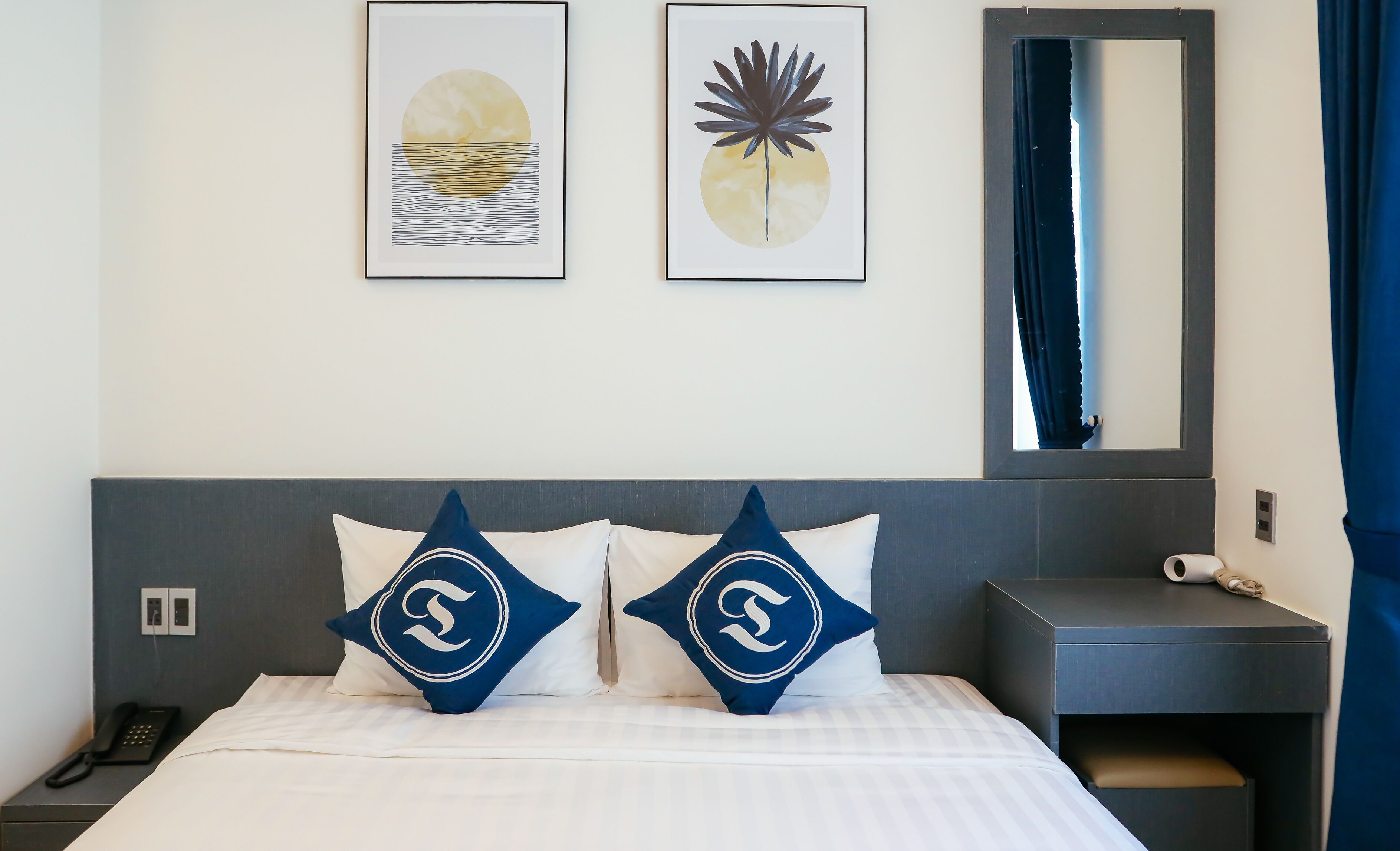 Habitación de hotel con cama, dos almohadas con un logotipo, teléfono en la mesita de noche, mesita de noche, espejo y dos obras de arte enmarcadas en la pared.