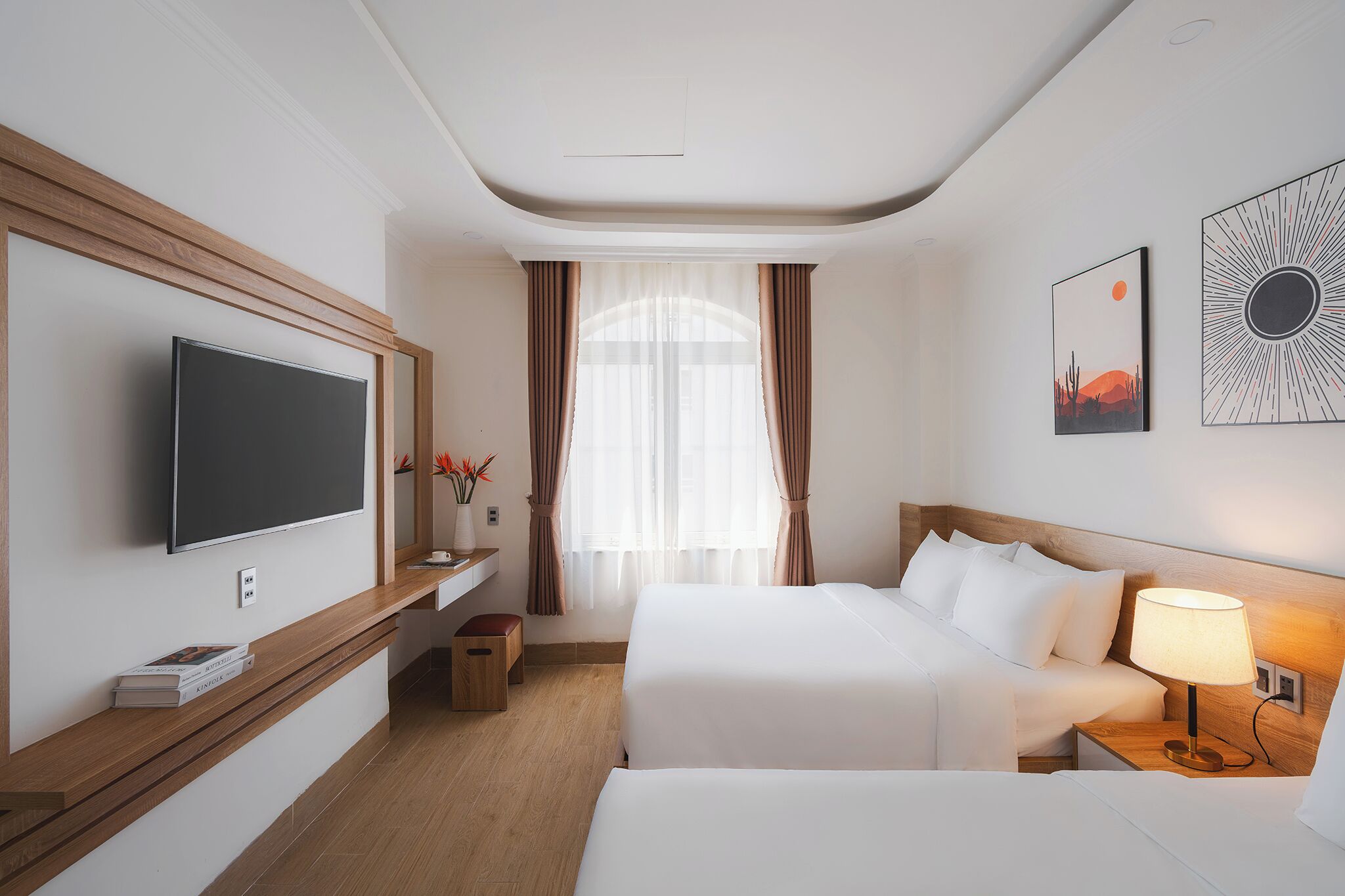A hotel room with a flat-screen TV, a bed, a bedside table with a lamp, a window with curtains, and artwork on the wall.