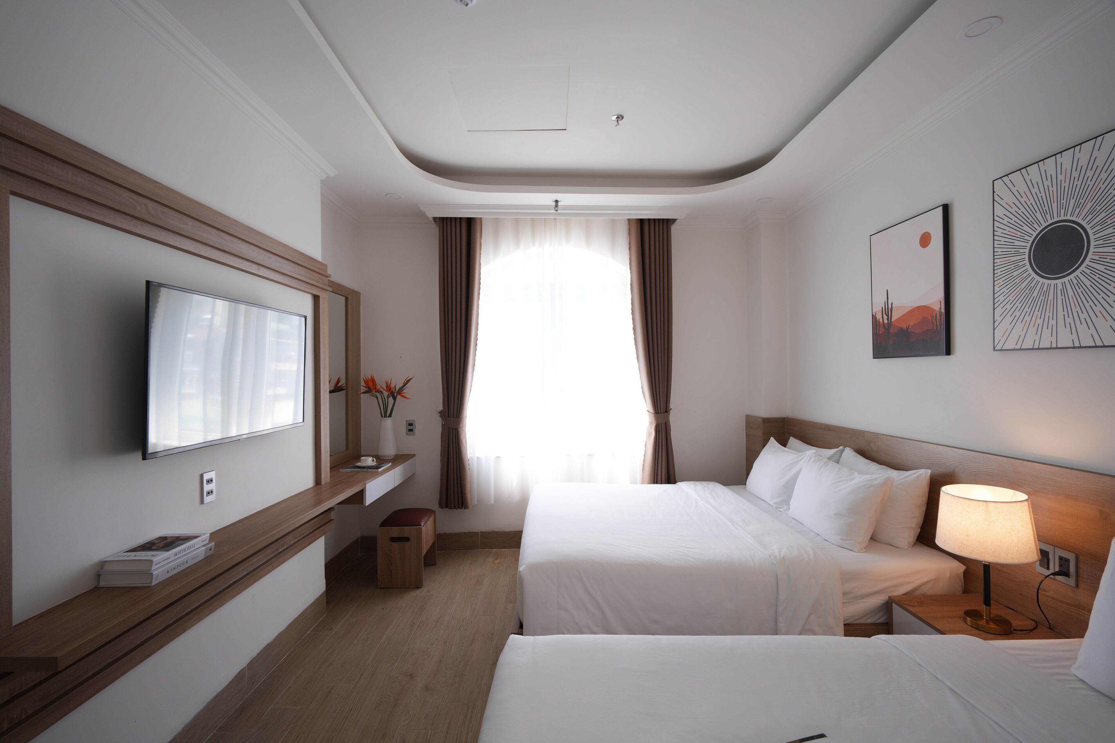 A hotel room with two beds, a TV mounted on the wall, a bedside table with a lamp, and a window with curtains.