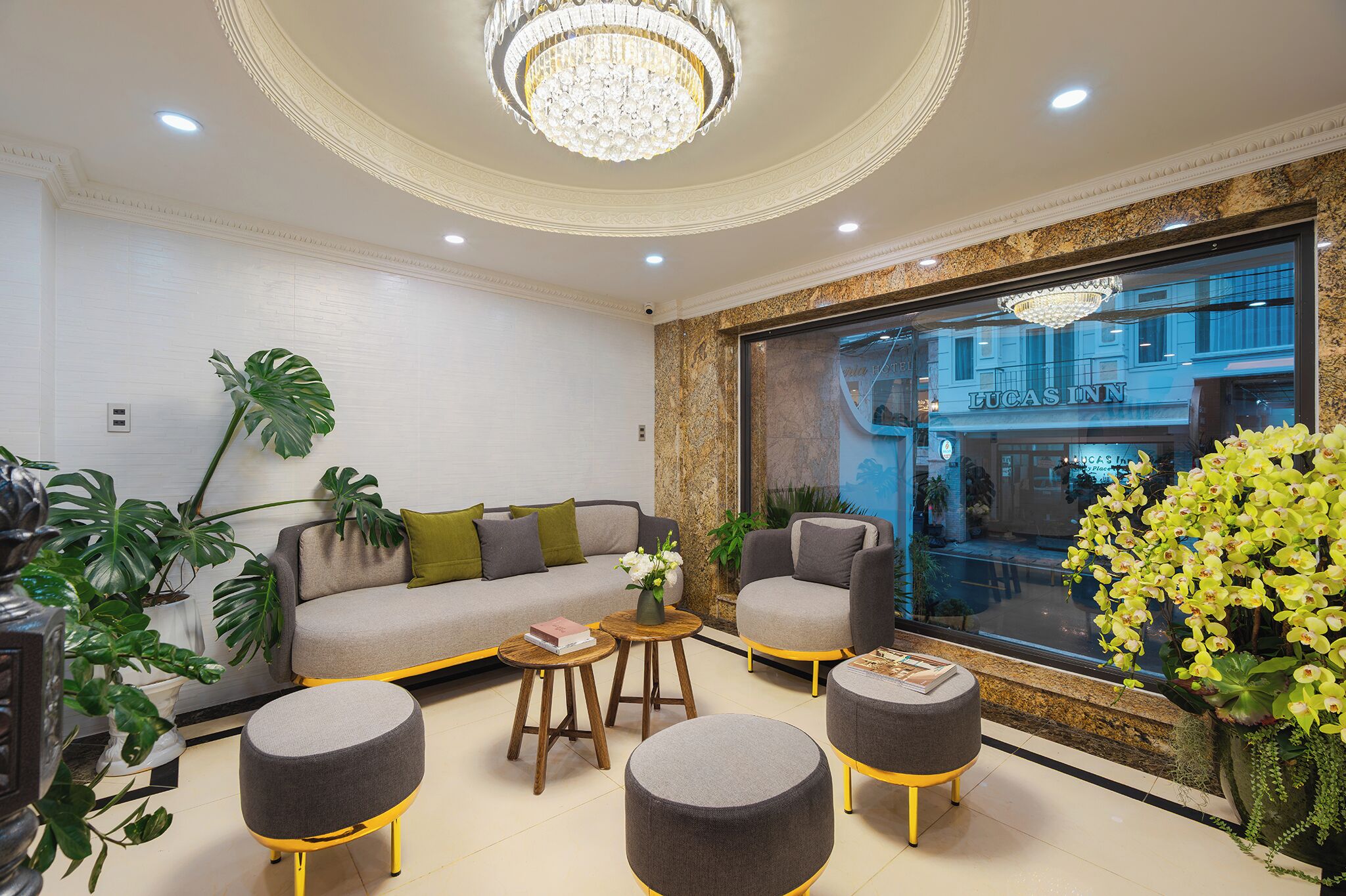 A modern living room with a sofa, ottomans, and a coffee table. There is a large window with a view of a building and a chandelier above.