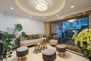 Una sala de estar moderna con un sofá, otomanas y una mesa de centro. Se ve una ventana grande con vistas a un edificio y un candelabro colgando.