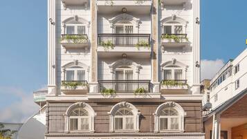 A multi-story hotel with a classic architectural design, featuring balconies with plants, a prominent entrance, and a clear sky above.