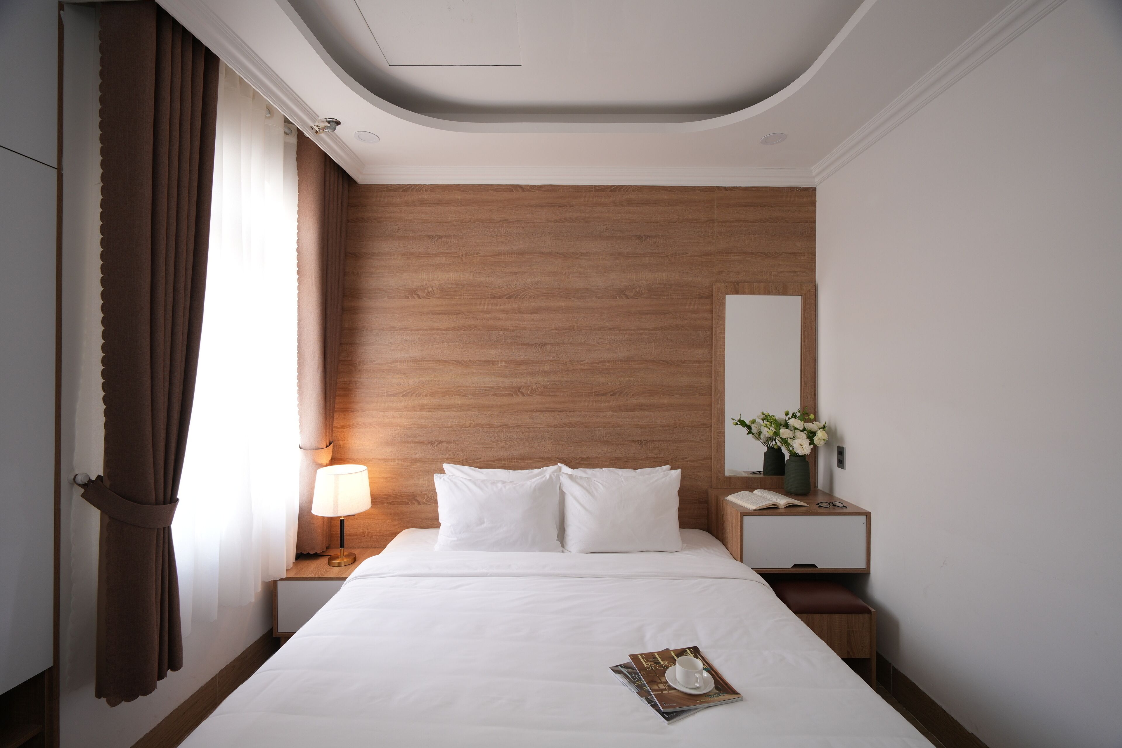 Une chambre d’hôtel avec une tête de lit en bois, une table de chevet avec une lampe, une petite table de nuit avec un vase de fleurs et un magazine sur le lit.