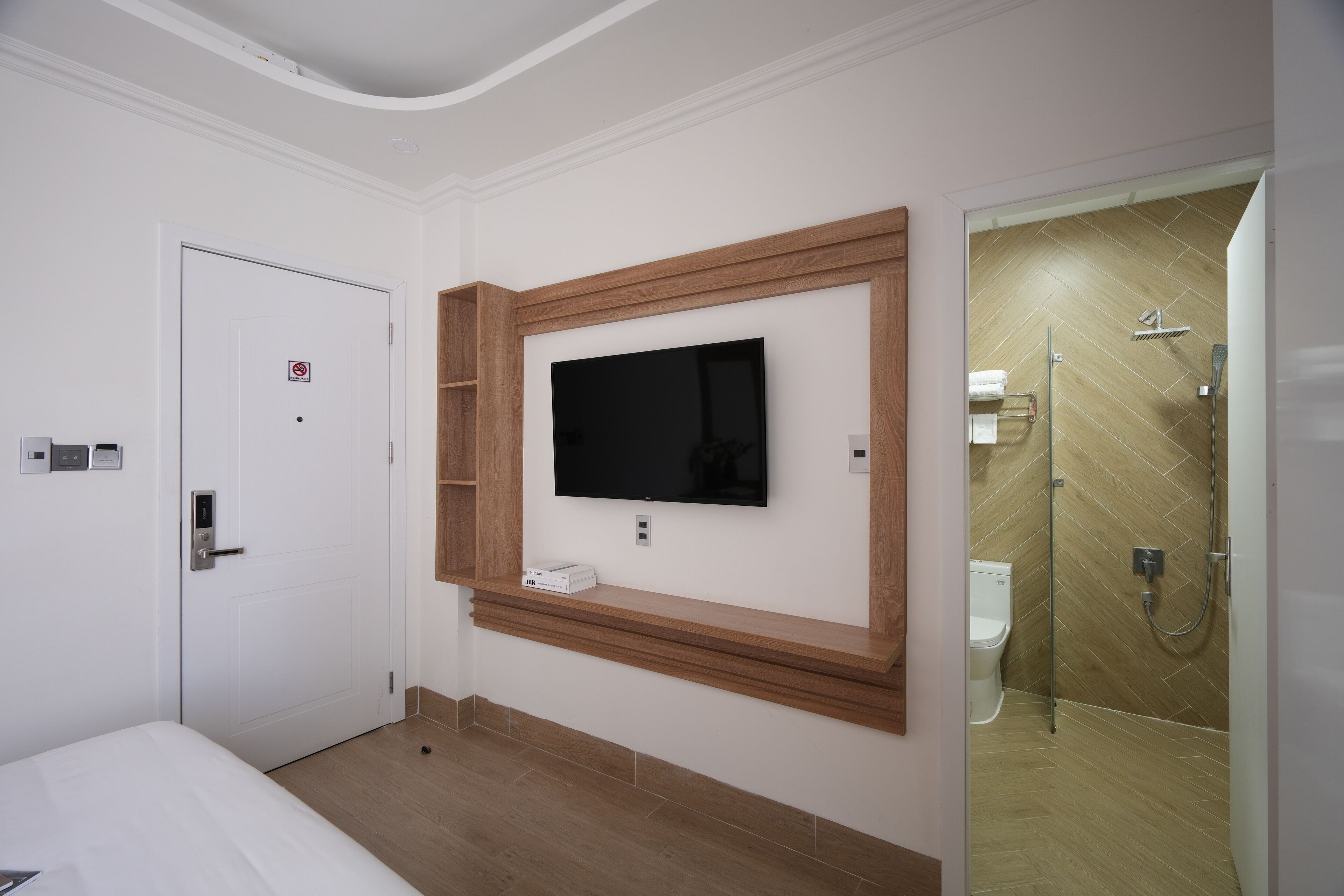 A hotel room with a bed, a mounted TV, a wooden shelf, a shower area, and a door with a no-smoking sign.